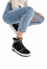 Persona con vaqueros de color azul claro y zapatillas altas negras con acentos metálicos, agachada contra un fondo blanco.