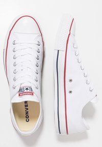 White canvas sneakers with red and navy blue striping, round rubber toe, silver eyelets, and white laces. "Converse All Star" label visible.