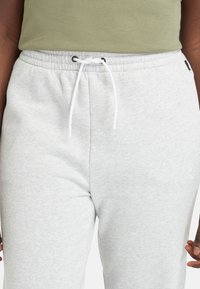 Pantalons de jogging en polaire gris clair avec une taille élastique et un cordon blanc. Présente une broderie de logo discrète sur la jambe inférieure. Texture douce.