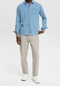 Camisa de denim azul claro con un bolsillo delantero, camiseta de manga larga gris debajo, combinada con pantalones beige y zapatillas blancas, de pie.