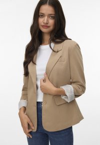 Blazer beige chiaro con chiusura a un pulsante, rever a scollatura, e dettagli a righe sui polsini, abbinato a una t-shirt bianca e jeans blu.