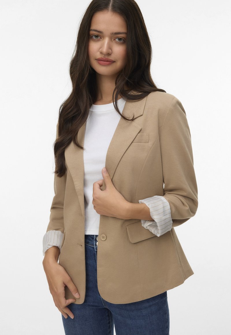 Blazer beige chiaro con chiusura a un pulsante, rever a scollatura, e dettagli a righe sui polsini, abbinato a una t-shirt bianca e jeans blu.