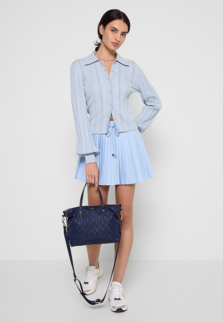 Himmelblauer Strickcardigan mit Kragen, kombiniert mit einem crémefarbenen, plissierten Rock. Das Model hält eine dunkelblaue Handtasche mit goldenen Beschlägen.