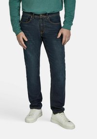 Donkerblauwe denim jeans met oranje stiksels, gecombineerd met witte sneakers. Lange mouwen groene top zichtbaar. Casual pasvorm.