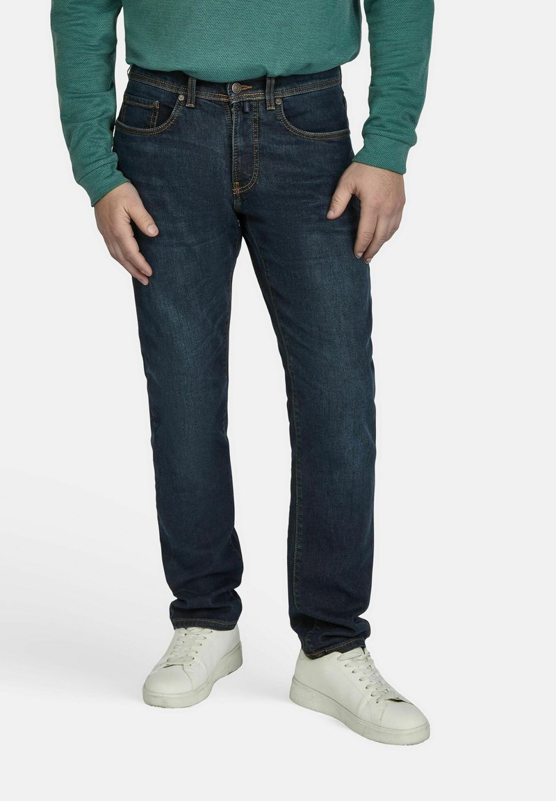Donkerblauwe denim jeans met oranje stiksels, gecombineerd met witte sneakers. Lange mouwen groene top zichtbaar. Casual pasvorm.