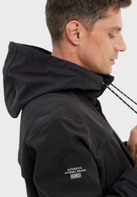 Veste à capuche noire avec un extérieur lisse, des cordons de serrage ajustables et un écusson logo sur la manche. Tissu texturé et design cintré.