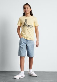Gul bomulds T-shirt med en sort grafik af en surikat med teksten "HVEM MIG?". Parret med lyseblå denimshorts og hvide sneakers.