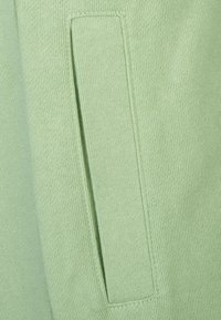 Hellgrüner Strickstoff mit seitlicher Tasche und rechteckigem Klappdeckel, glatte Textur und saubere Nahtdetails.