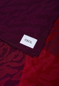 Struktureret stof i dyb lilla og rød med et mønstret design. Mærket læser "CECIL." Kanterne er flossede for et afslappet udtryk.