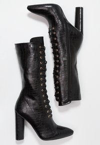 Bottes à talons hauts noires en similicuir texturé. Elles présentent un laçage devant avec des œillets dorés et une fermeture éclair latérale.