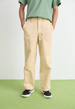 Beige, loose-fit trousers made of cotton with straight legs, featuring side pockets and a button closure. Worn with black sneakers.