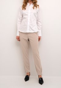 Bílá košile s dlouhým rukávem a knoflíky, spárovaná s béžovými zúženými kalhotami se zipy na kotnících a černými špičatými botami.