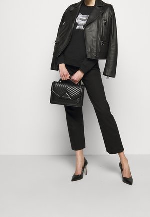 Woman wearing black leather jacket over black sweatshirt and pants, holding black quilted handbag, wearing black high heels, standing on grey floor.