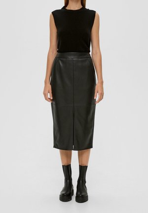 Vrouw draagt mouwloze zwarte top, zwarte leren midi-rok met hoge taille en split vooraan, en zwarte enkellaarsjes, staand tegen een effen achtergrond.