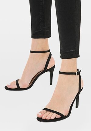 Sandales noires à talons hauts avec fines brides à la cheville et aux orteils, portées avec un pantalon noir court sur fond blanc uni.