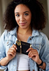 Černá peněženka se zlatými doplňky, která má střapec a logo značky. Texturovaný povrch má jemný vzor. Džínová bunda přes bílé tričko.