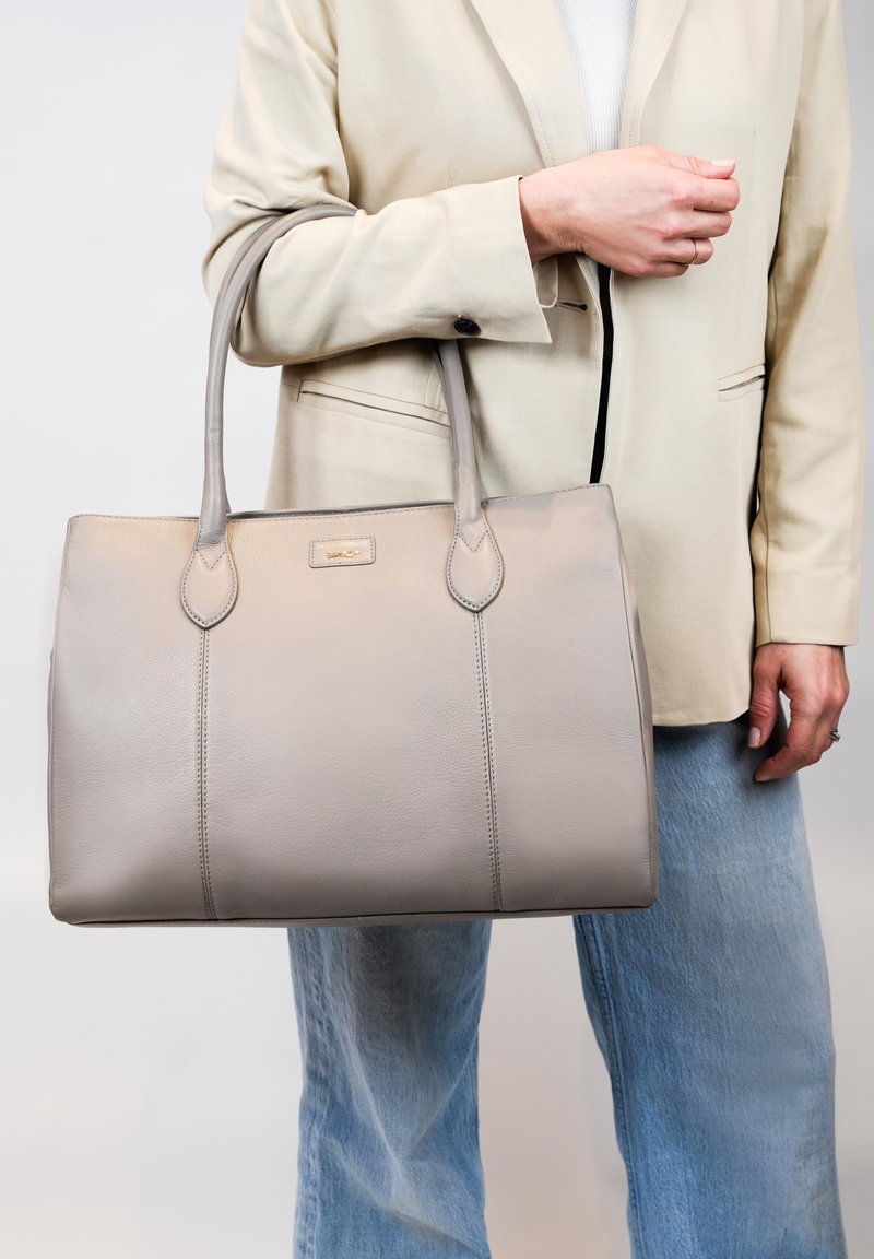 Grijze leren handtas met twee handvatten, gestructureerde vorm en subtiele stikdetails. Draagt met een crème blazer en lichtblauwe spijkerbroek.