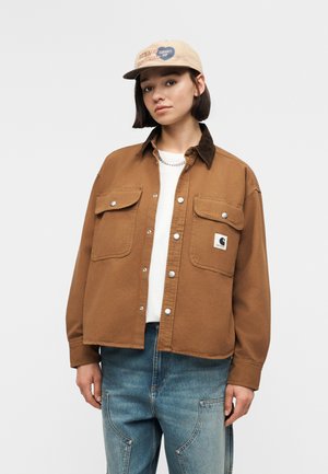 Fiatal személy világosbarna sapkával, barna, gombos, első zsebes dzsekivel, fehér inggel és kék farmerrel, egyszerű háttér előtt.