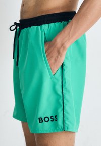Tirkizne kupaće hlače s crnim elastičnim pojasom i vezicom. Ima bočne džepove i 'BOSS' logo u crnoj boji na donjem lijevom dijelu.
