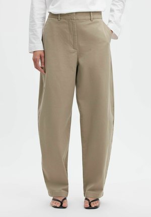 Personne portant un pantalon beige à jambes larges et des sandales noires, avec une main dans la poche et une chemise blanche à manches longues.