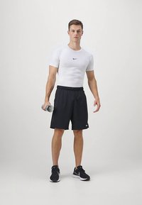 Vit kortärmad skjorta med en svart Nike-logotyp, svarta shorts och svarta träningarskor, som håller en silver hantel. Neutral bakgrund.