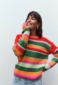 Stickad tröja med flerfärgade ränder i röd, grön, orange, rosa och kräm. Har ribbade ärmslut och nederkant. Bärs med ljusblå jeans.