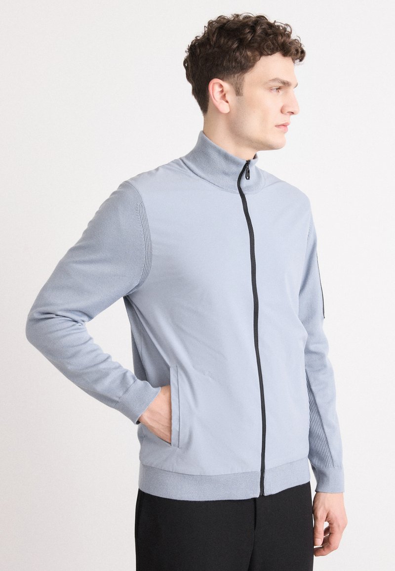 Šviesiai mėlynas zip-up švarkas su aukštu apykakle, briaunuota tekstūra ant pečių ir šoninėmis kišenėmis. Juodi užtrauktukai ir prigludę rankogaliai.
