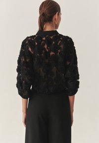 Top trasparente nero con applicazioni di rose 3D, dotato di colletto e maniche a tre quarti, abbinato a pantaloni neri su misura.