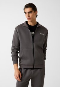 Giacca grigia con zip e colletto alla coreana, con strisce nere laterali e logo "GUESS" sul petto. Realizzata in tessuto liscio e morbido.