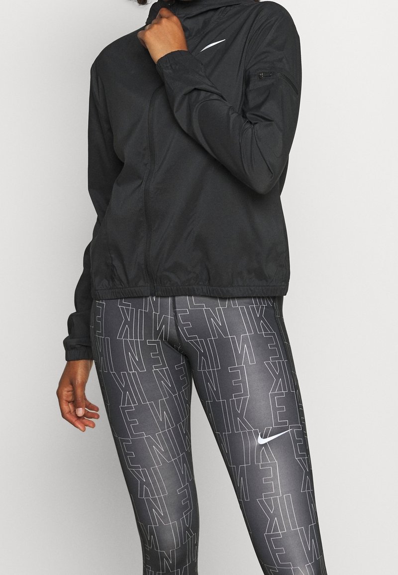 Veste légère noire avec fermeture éclair à l'avant et capuche, associée à un legging gris présentant un motif en répétition du logo "Nike" en blanc.