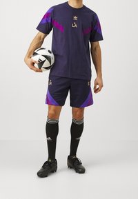 Marinträna Adidas sportset i marinblått med lila detaljer; t-shirt, shorts, svarta knästrumpor och svarta fotbollsskor. Håller i en fotboll.