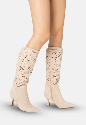 Beige ruskind støvler til knæet med laserudskårne blomster mønstre, spidse tæer, slanke hæle og sidelommer, båret på krydsede ben.