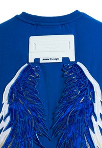 Camisa de algodón azul con diseños de alas bordados en blanco y azul en la espalda, con una etiqueta blanca que muestra "MWM Design".