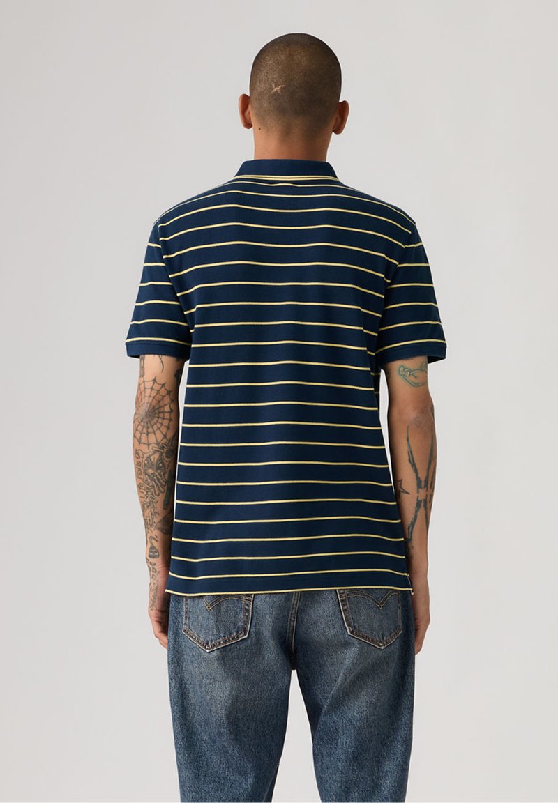Mand med tatoveringer iført en marineblå poloshirt med gule striber og blå jeans, stående med ryggen mod en ensfarvet baggrund.