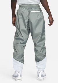 Calças de treino cinza em nylon com cintura elástica, detalhes brancos nos punhos e um bolso traseiro com fecho éclair.