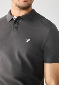 Sort bomulds-polo shirt med krave med to knapper, der har et hvidt broderet fuglelogo på venstre bryst. Korte ærmer med ribbede manchetter.