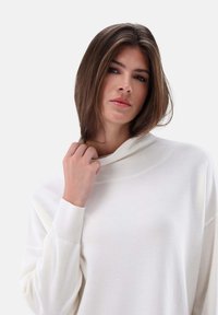 Weißer, langärmeliger Pullover mit lockerer Passform und hohem Kragen. Das Material wirkt weich und strukturiert, mit Rippen an den Bündchen.