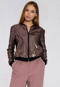 Błyszcząca kurtka bomberka w kolorze brązowym z czarnym zamkiem i prążkowanymi mankietami. Połączona z jasnoróżowymi spodniami o luźnym kroju.