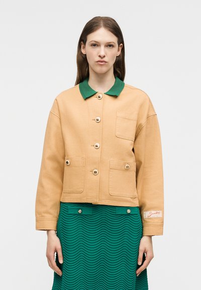 Γυναίκα με μπεζ σακάκι με κουμπιά και μπροστινές τσέπες, και πράσινο πουκάμισο με γιακά κάτω από πράσινη υφασμάτινη φούστα.
