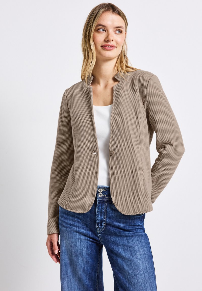 Blazer beige testurizzato con scollo rotondo e chiusura a bottone singolo, abbinato a una maglietta bianca e jeans a gamba larga blu.
