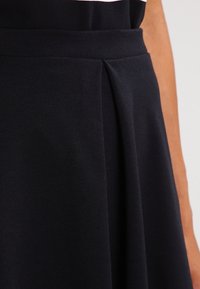 Svart plisserad tyg med en slät textur och en detalj vid midjan. Nära bilden visar fall och konstruktion av plagget.