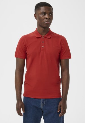Junger Mann trägt ein rotes Poloshirt mit kurzen Ärmeln und blaue Jeans, steht vor einem schlichten weißen Hintergrund.