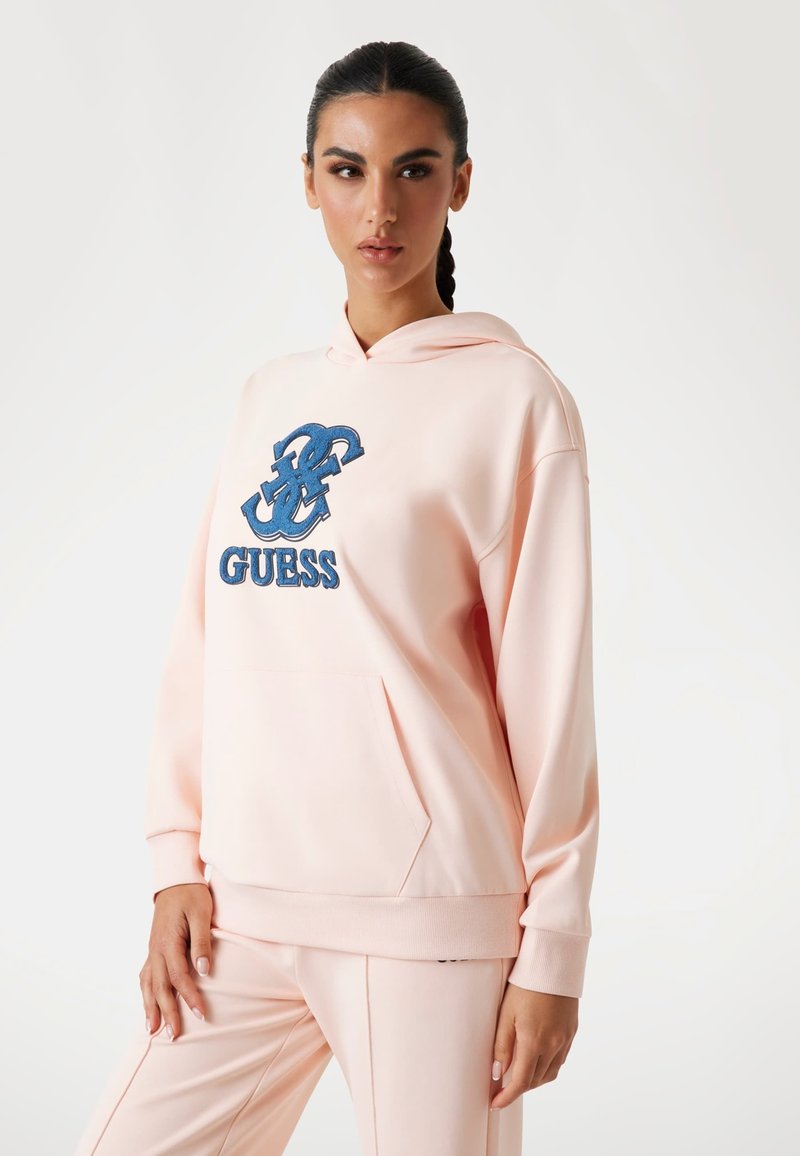 Sudadera rosa claro con bolsillo frontal, logo bordado azul "GUESS" y diseño decorativo. Tela suave, ajuste relajado, puños acanalados.