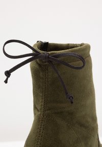 Gros plan sur une botte en daim vert olive avec un lacet noir noué en un nœud près de son bord supérieur.