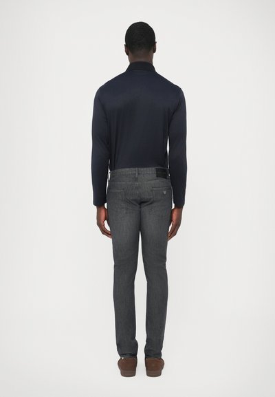 Tmavomodrá polokošeľa s dlhým rukávom v kombinácii s tmavosivými džínsami rovného strihu a hnedými topánkami, zobrazená zozadu na jednoduchom bielom pozadí.