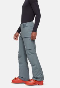 Pantalon de ski gris à coupe ample, doté de poches zippées sur les côtés et d'un patch logo. Le mannequin porte des chaussures de ski orange vif pour le contraste.