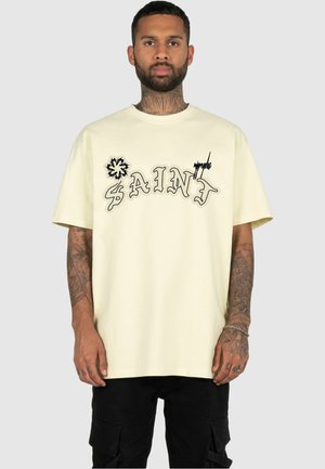 T-shirt in cotone color crema con testo stilizzato "SAINT" con contorno nero e grafica decorativa, maniche corte e vestibilità ampia.