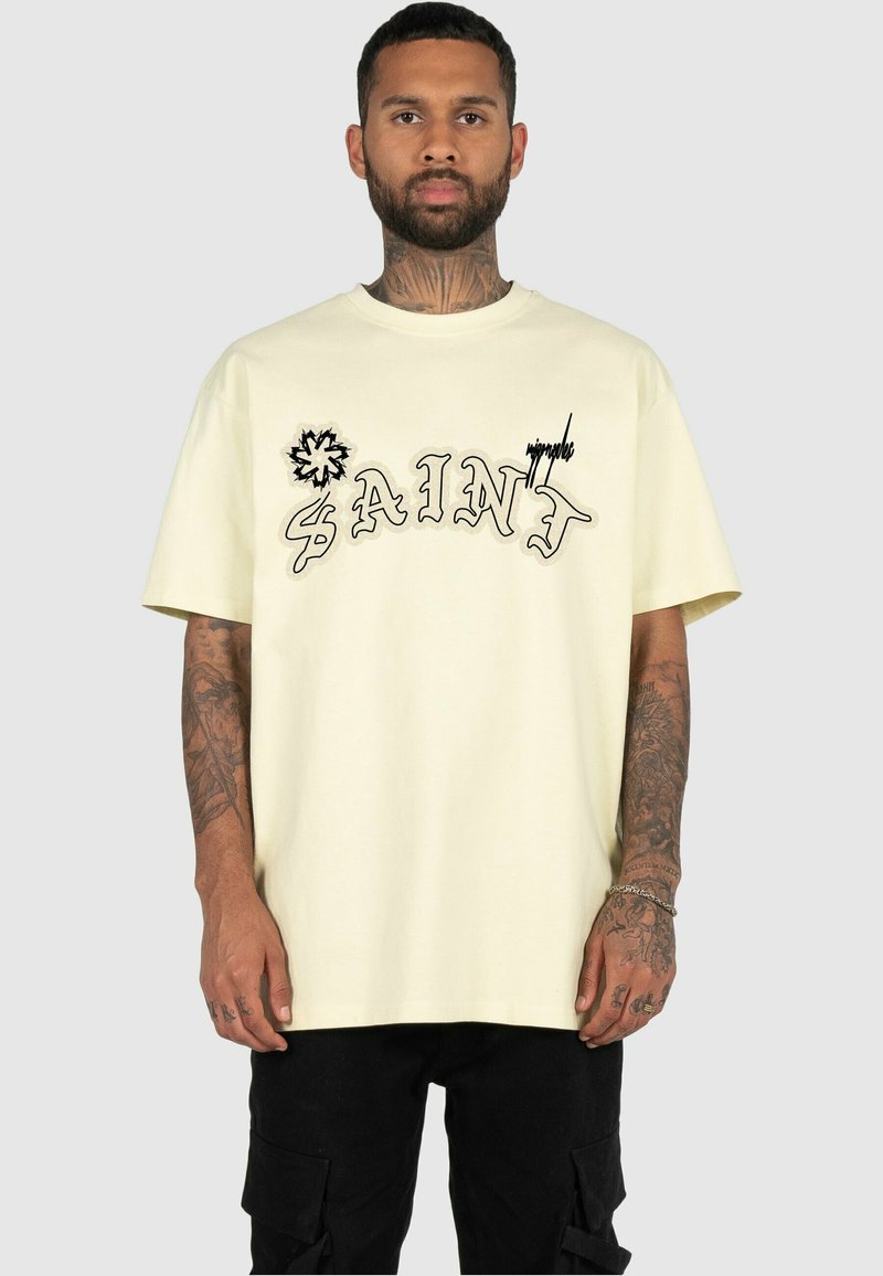 Kremowa bawełniana koszulka z krótkim rękawem, z projektu stylizowanego napisu "SAINT" w czarnym konturze oraz dekoracyjną grafiką, w luźnym kroju.