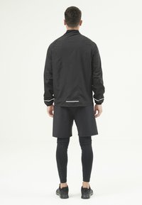 Homme debout de dos portant une veste noire, un short noir par-dessus un legging noir, et des chaussures de sport noires sur fond blanc.