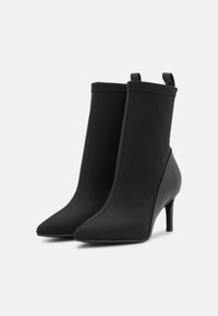 Ett par svarta ankelstövlar med spetsiga tår, smala klackar och dragflikar på vit bakgrund.